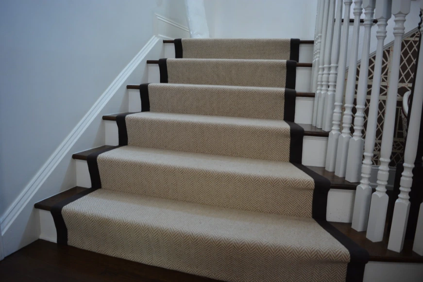 Herringbone pattern wala carpet runner jis ke dono taraf kaala (black) border hai, jo safed aur lakdi ki seedhiyon per laga hai.