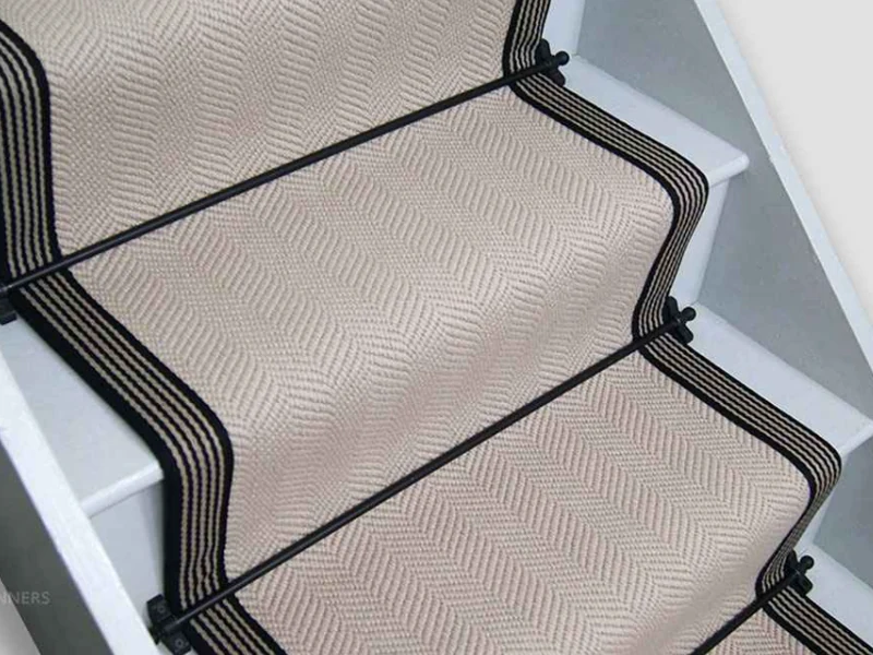 herringbone-patterned stair runner with a thin black and beige striped border, secured to white wooden stairs with black metal stair rods.