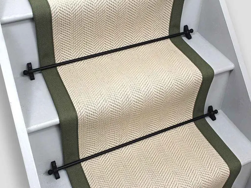 herringbone wool stair runner with olive green fabric borders, secured to white wooden stairs by black metal stair rods.