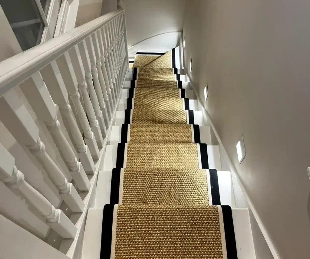 stairs are painted white, and the natural-fiber runner is a golden-tan color with a textured, woven pattern.